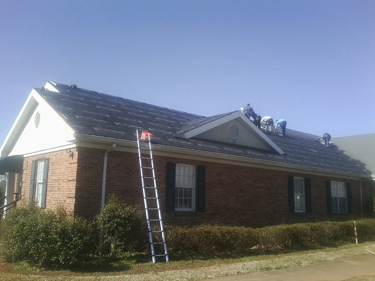 Crew tearing shingles off a brick ranch roof with a ladder set — Metal Roof Repair work by our crew in New Waverly