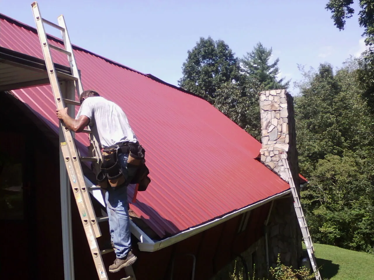 Roofer on a ladder beside a red metal roof and stone chimney — Soffit & Fascia Repair by Roofing Brothers in New Waverly